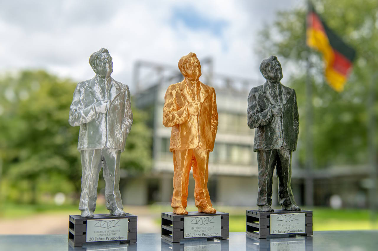 Pokale von Helmut Schmidt in den Farben silber, bronze und dunkelgran (von links nach recht) sowie dem Hauptgebäude der Helmut-Schmidt-Universität und der Deutschlandflagge im Hintergrund